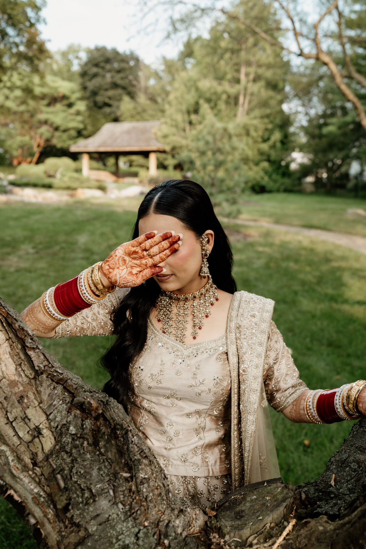 Bride in a gold embroidered lehenga covering her eyes with a hennaed hand, red chooda bangles on her wrists