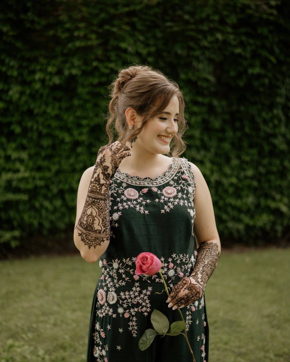 Smiling bride in a dark green embroidered outfit with full mehndi on her hands, holding a pink rose