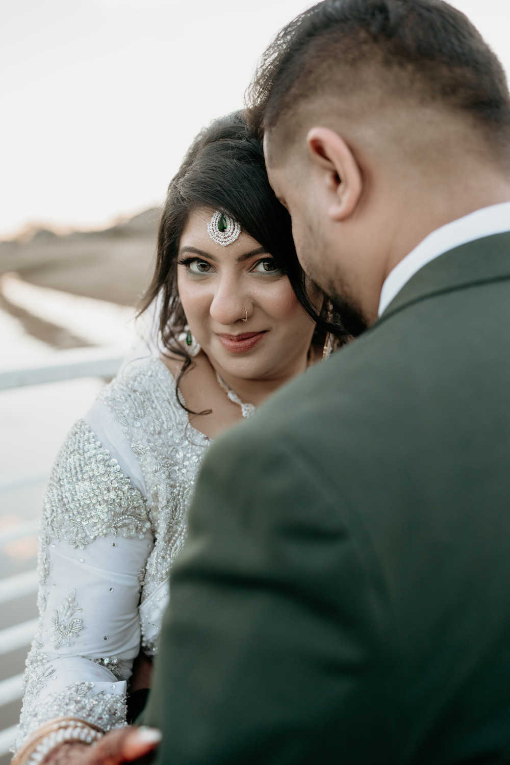 Bride in a white embellished outfit and emerald maang tikka smiling at the camera as the groom in a dark green suit leans into her