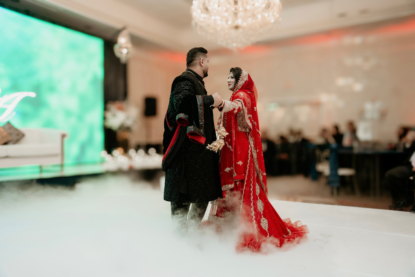 Bride in a red embroidered outfit and groom in a black sherwani dancing on a cloud of low fog beneath a crystal chandelier