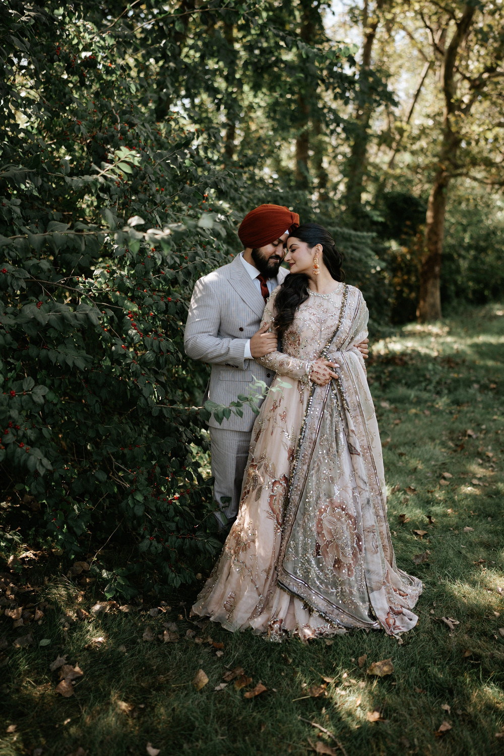 Groom in a grey pinstripe suit and rust turban holding his bride in a blush embellished lehenga among green trees