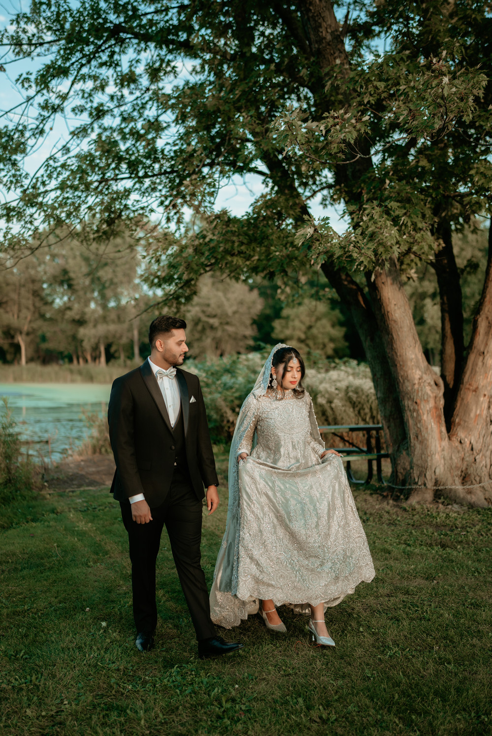 Bride lifting her silver embroidered gown as she walks beside the groom in a black tuxedo under a large tree by a lake