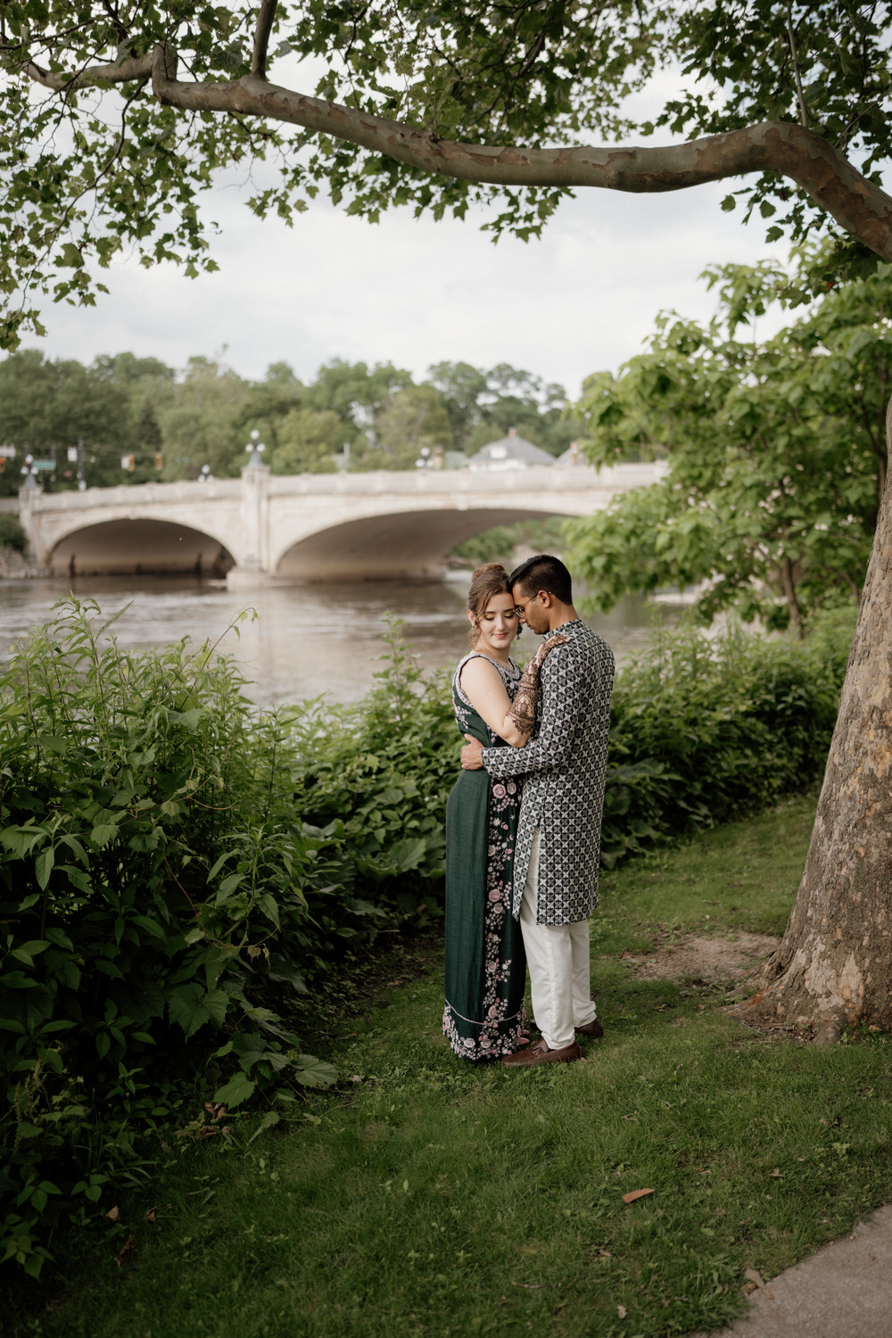 Couple embracing by a river under a sycamore branch, with a stone arch bridge behind them