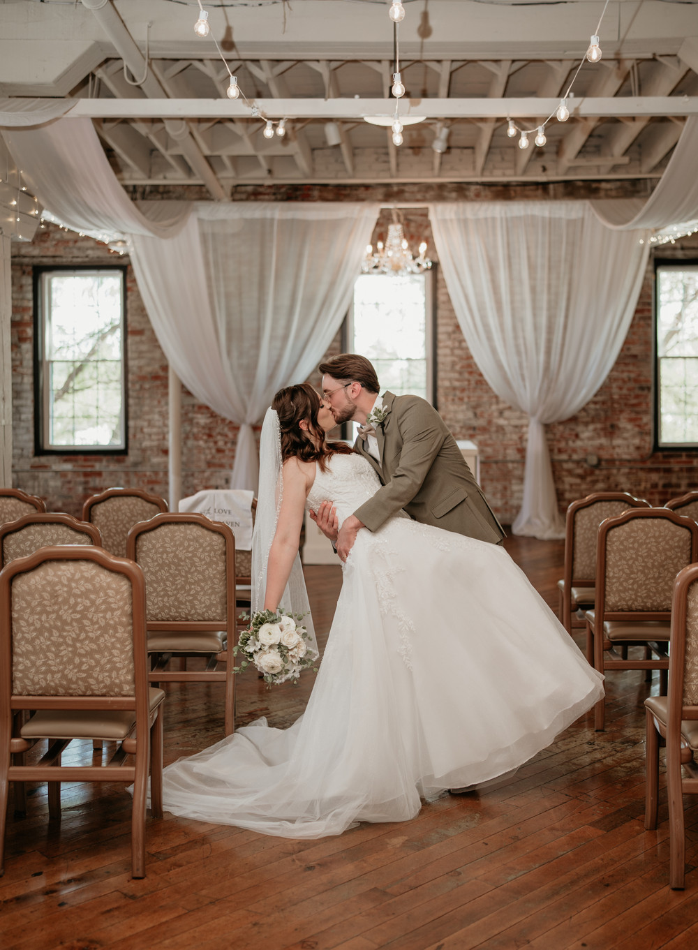Groom in a taupe suit dipping and kissing his bride in a white gown in an aisle of chairs beneath string lights