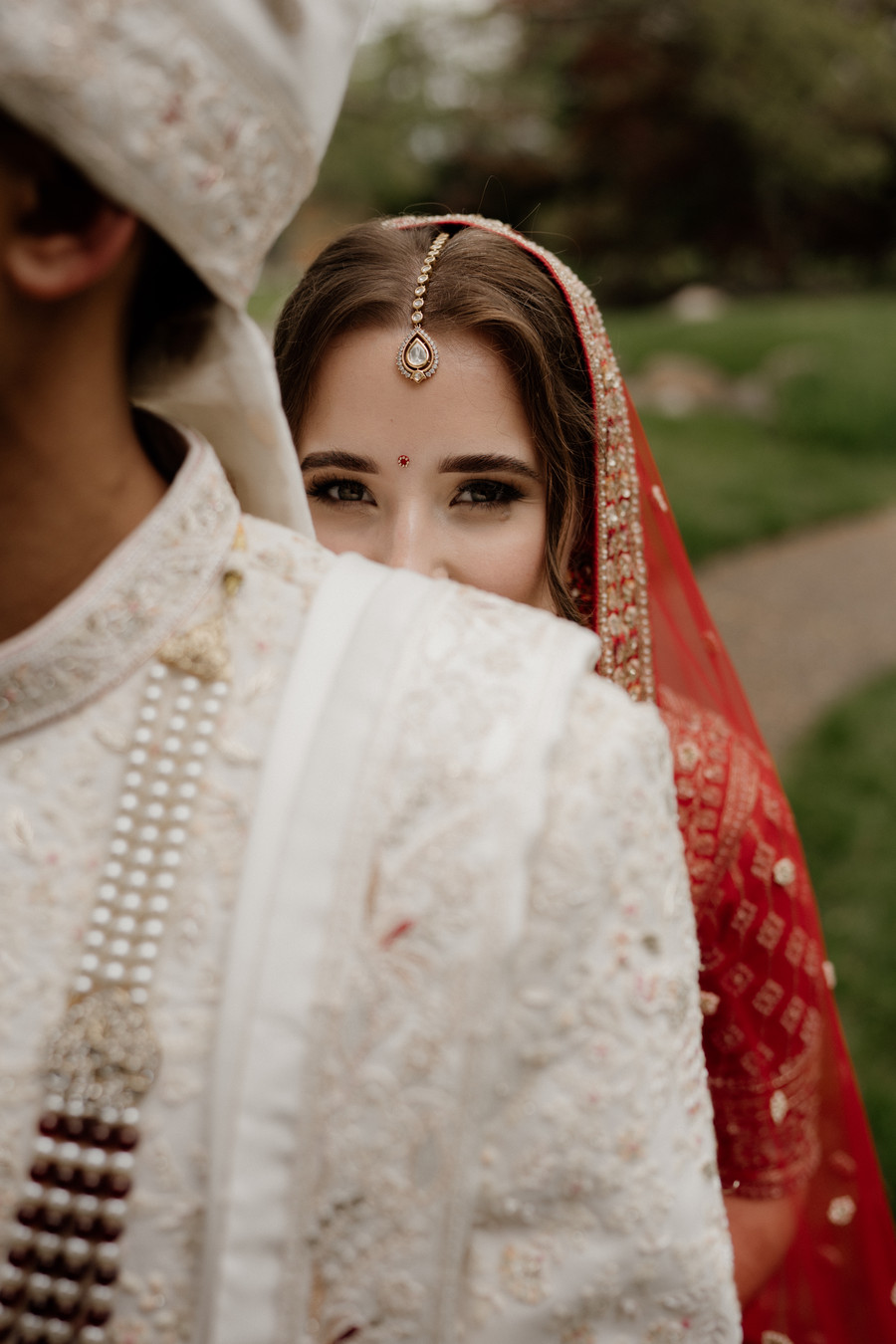 Bride in a red dupatta and maang tikka peeking over the shoulder of the groom in an embroidered ivory sherwani